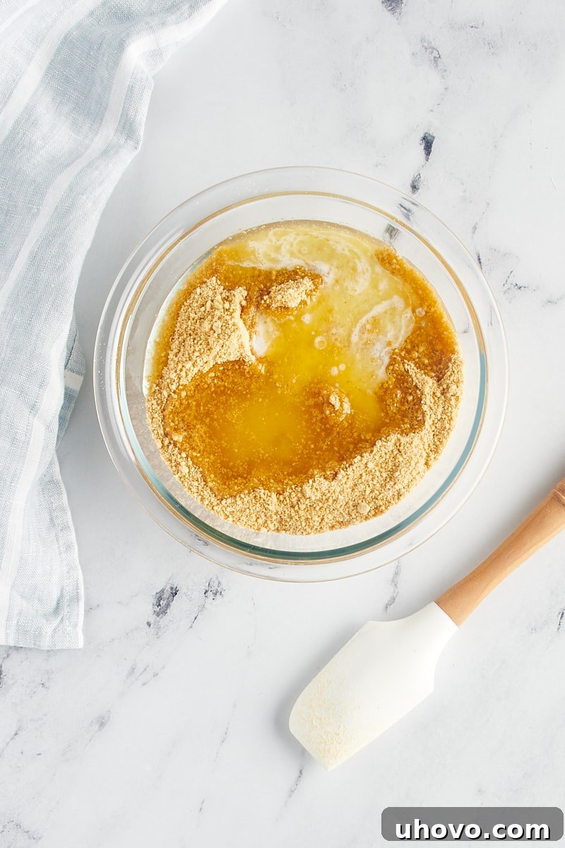 Melted butter being poured into a bowl containing graham cracker crumbs and sugar, ready for mixing.