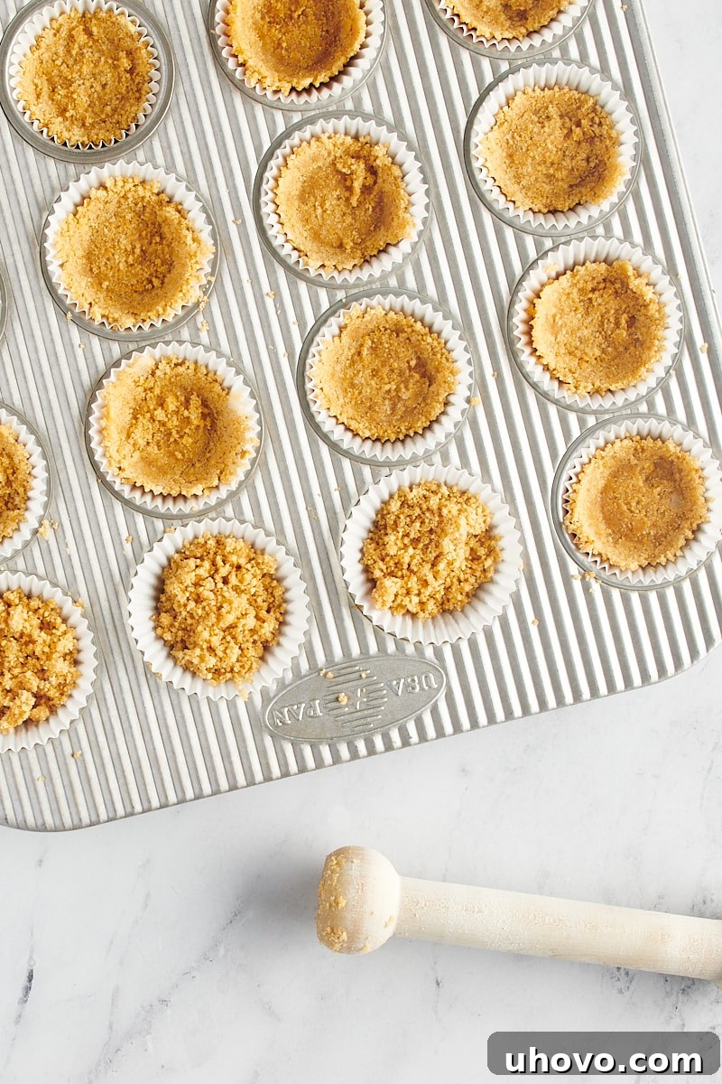 Evenly portioned graham cracker crust mixture settled into the bottom of a mini muffin tin lined with paper cups.