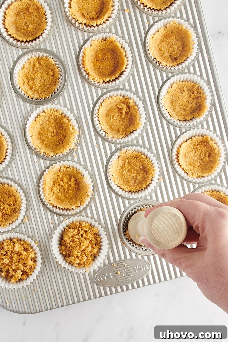 A tart tamper gently pressing the graham cracker crust into the bottom of a lined mini muffin cup, ensuring a firm base.