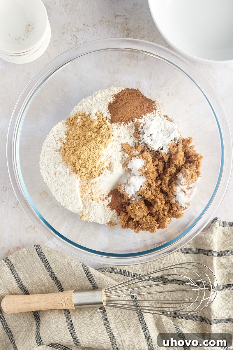 An overhead shot of various dry ingredients for gingerbread muffins – flour, brown sugar, baking powder, baking soda, ginger, cinnamon, nutmeg, and salt – meticulously arranged in a clear glass mixing bowl.