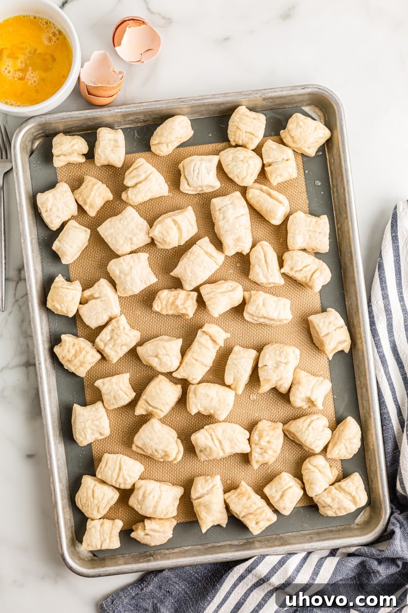 Pretzel dough pieces, fresh from the baking soda bath, arranged on a baking sheet lined with a silicone mat, ready for their egg wash.