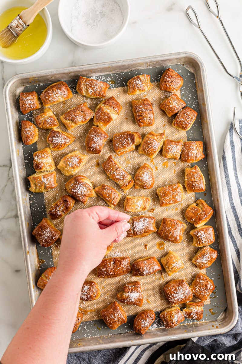 Coarse salt being sprinkled over hot, buttered pretzel bites on a baking sheet, adding the classic pretzel topping.