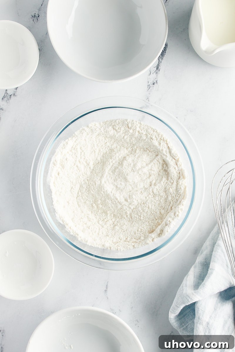 A glass mixing bowl holds whisked dry ingredients for the cake: flour, sugar, baking powder, and salt.