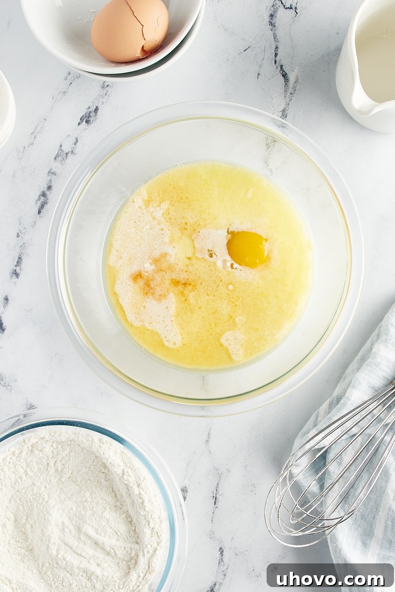 A separate glass mixing bowl contains the wet ingredients: egg, milk, slightly cooled melted butter, and vanilla extract, all whisked together.