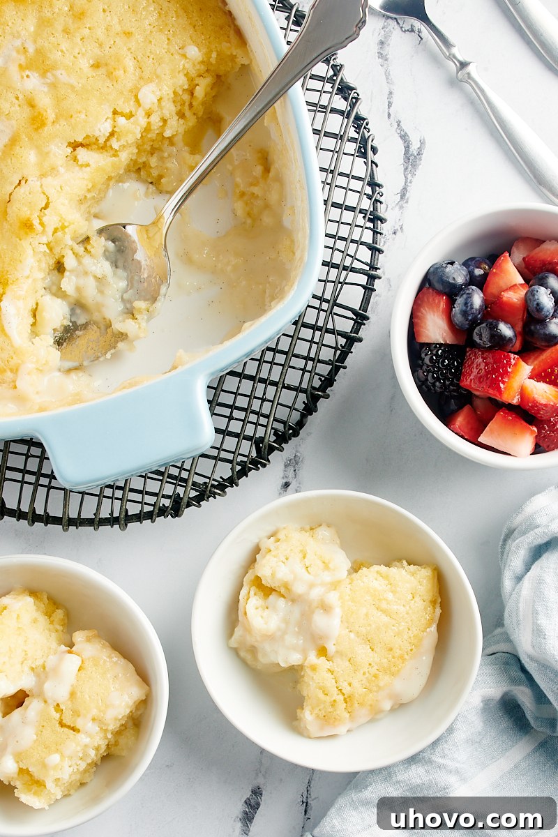 An artfully composed overhead shot displaying two plated servings of Vanilla Pudding Cake, the remainder of the cake in its pan, and a side bowl brimming with fresh, colorful mixed berries, ready for garnishing.