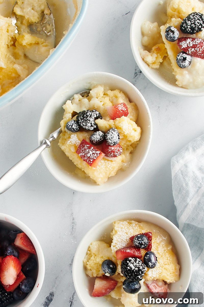 A stunning overhead perspective of three elegant servings of Vanilla Pudding Cake, each nestled in a white bowl and beautifully adorned with fresh mixed berries and a delicate dusting of confectioners' sugar.