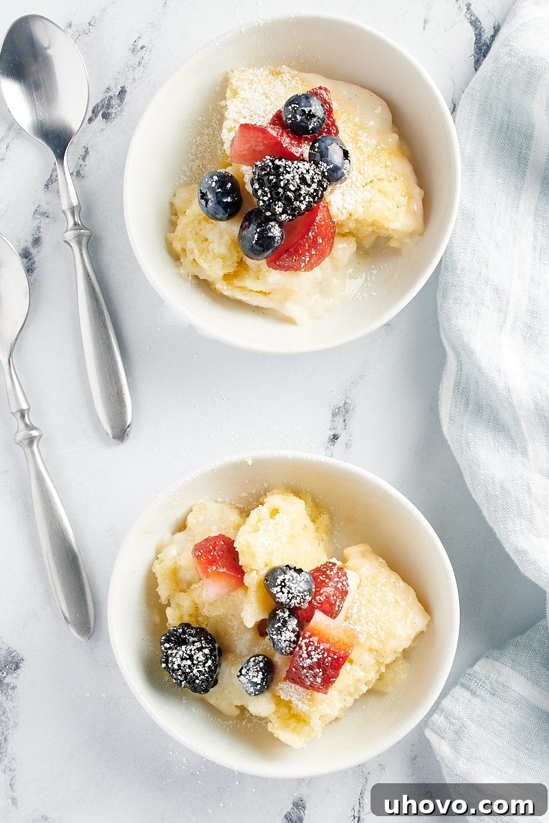 An inviting overhead shot showcasing two servings of warm Vanilla Pudding Cake in pristine white bowls, each generously topped with an assortment of fresh, vibrant berries.