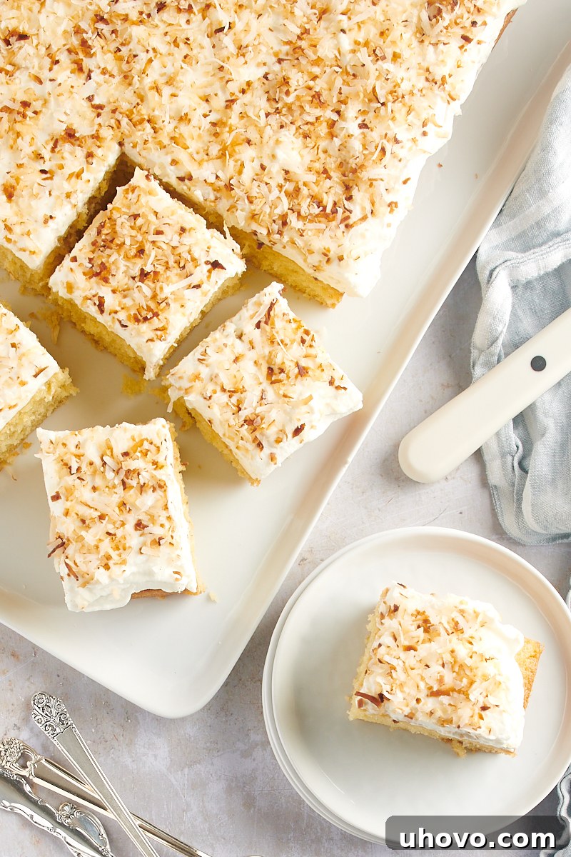 overhead view of Coconut Sheet Cake on a white serving tray and a slice on a white plate, showcasing its fluffy frosting and toasted coconut garnish
