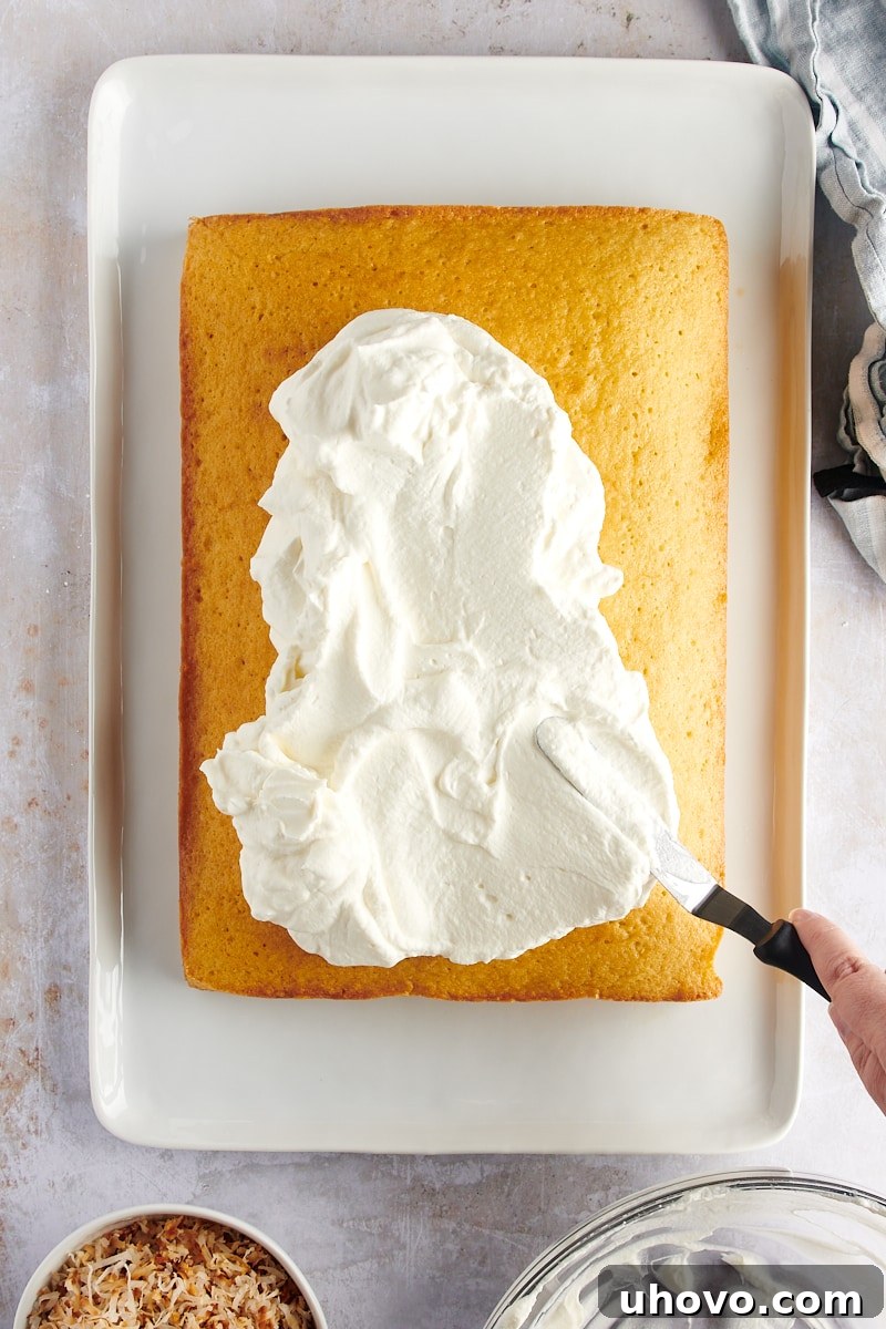 overhead view of a baker spreading whipped cream frosting evenly over the cooled coconut cake