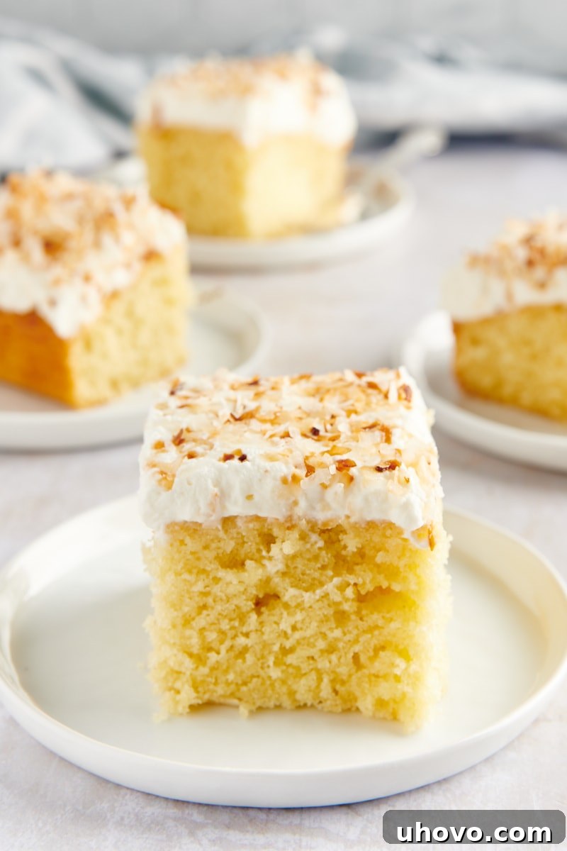 slice of Coconut Sheet Cake on a white plate with more slices on plates in the background, inviting viewers to enjoy