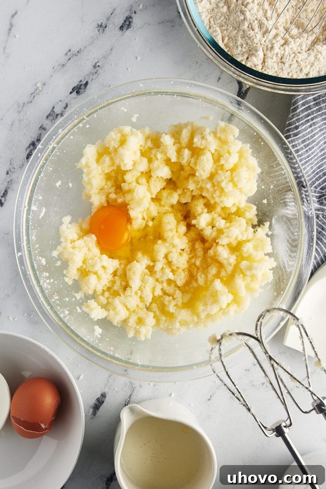 Overhead view of a large egg and pure vanilla extract added to the creamed butter and sugar mixture in a glass mixing bowl.