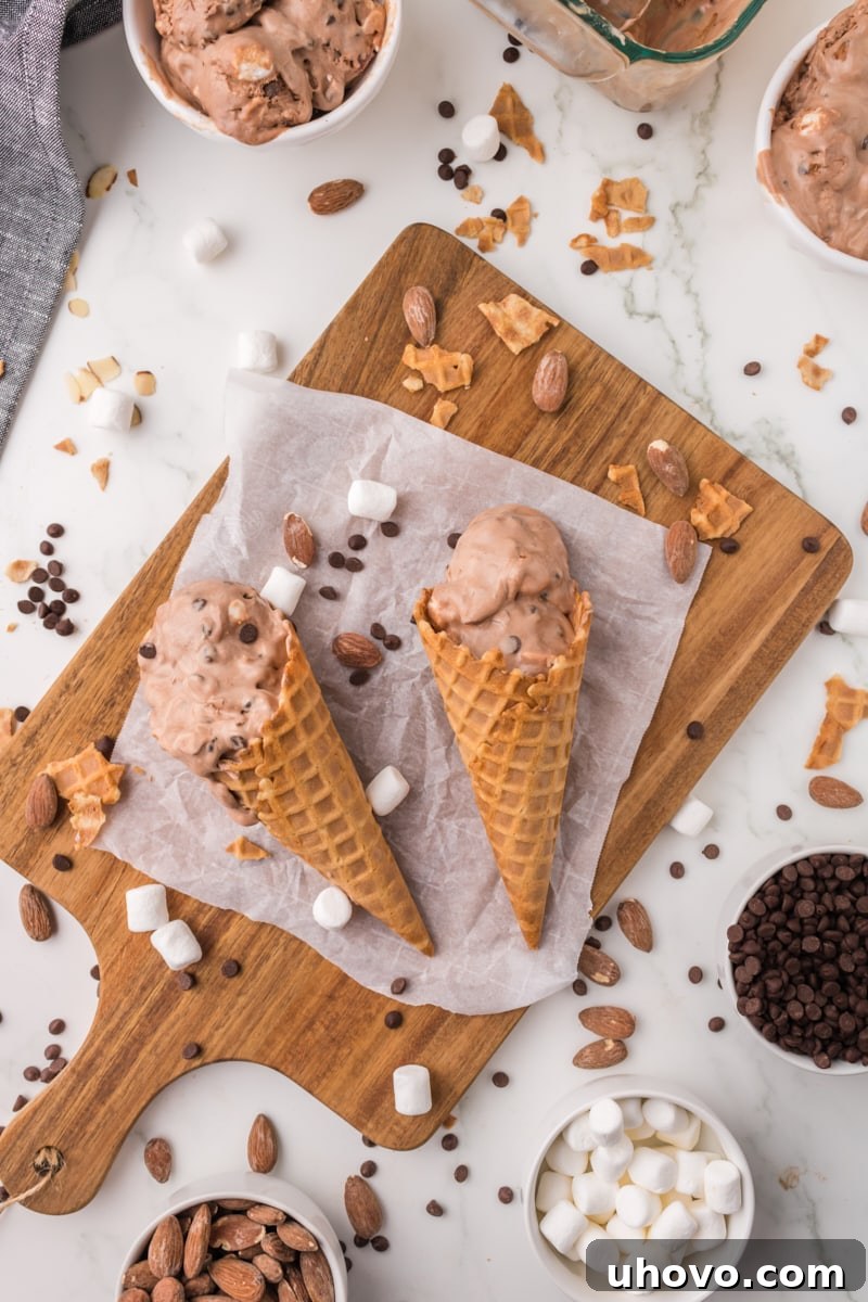 An overhead view of two waffle cones generously filled with Rocky Road ice cream, surrounded by scattered mini chocolate chips, marshmallows, and almonds for a tempting display.