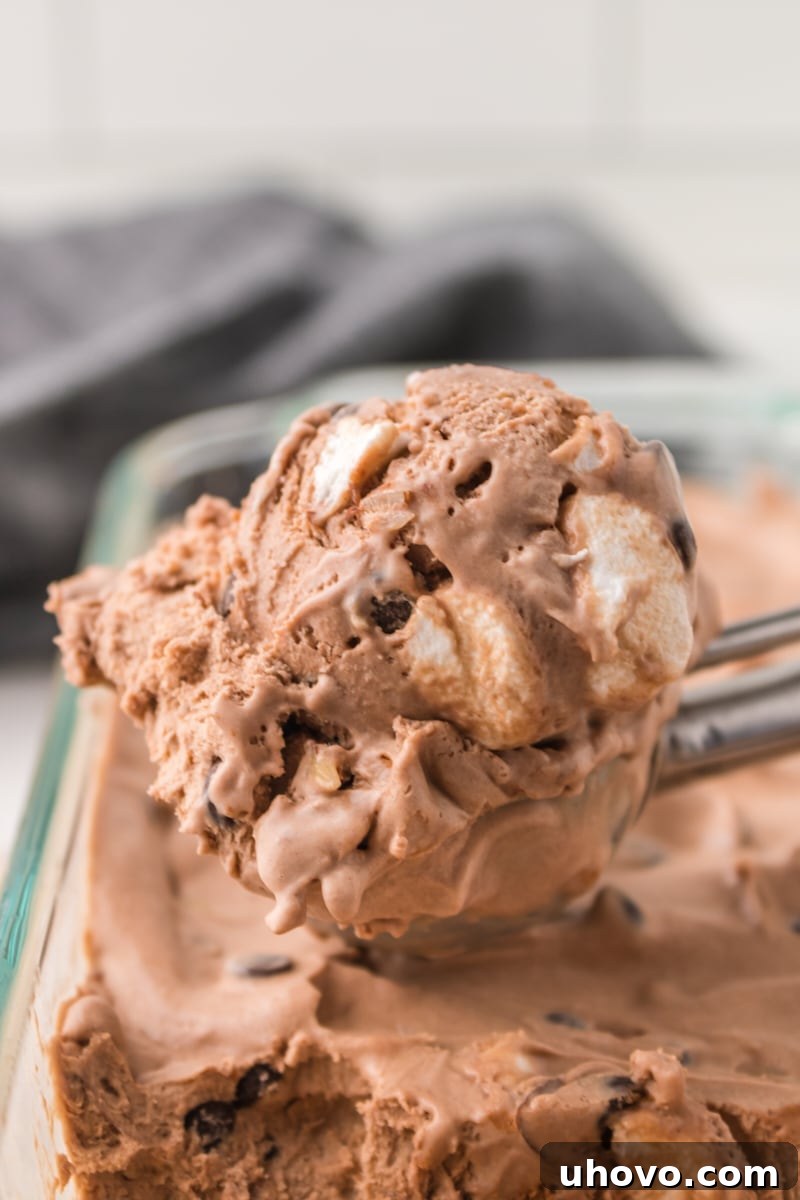 An ice cream scoop filled with freshly made Rocky Road Ice Cream rests on top of more ice cream in a clear glass pan, ready for serving.