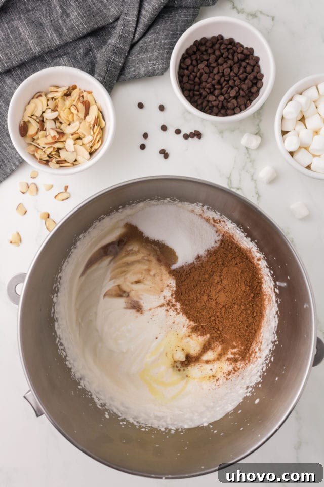 An overhead view showing cocoa powder, granulated sugar, vanilla extract, and almond extract being added to partially whipped heavy cream in a shining metal mixing bowl, forming the ice cream base.