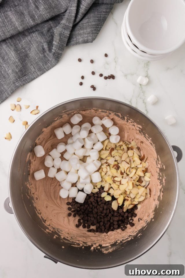 An overhead shot showcasing mini marshmallows, chocolate chips, and sliced almonds being carefully added to the freshly made chocolate ice cream mixture in a metal mixing bowl.