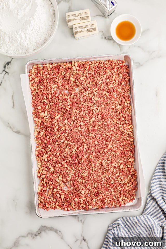 Overhead view of the strawberry crunch bits spread in a single layer on a parchment-lined sheet pan, allowing them to dry slightly.