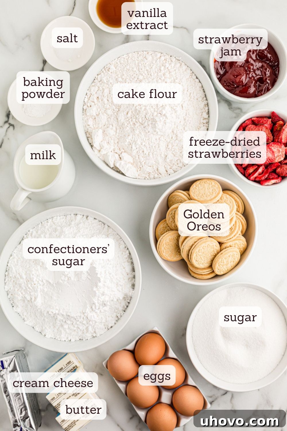 Overhead view of various ingredients laid out for strawberry crunch cake, including flour, sugar, butter, eggs, milk, jam, cream cheese, Golden Oreos, and freeze-dried strawberries.