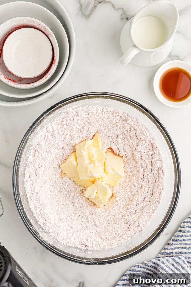 Overhead view of softened butter being added to the dry ingredients in a mixing bowl, prior to being mixed into a sandy texture.