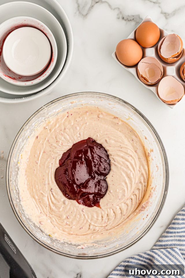 Overhead view of strawberry jam being added to a bowl of cake batter, the final ingredient before baking.