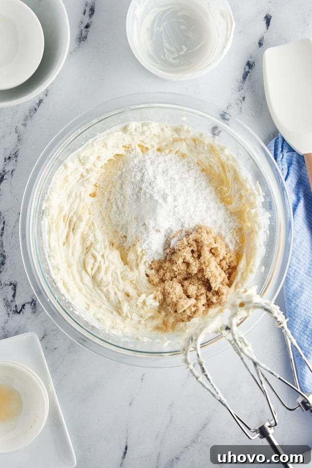 A close-up, overhead view of the mixing bowl where confectioners' sugar and brown sugar are being carefully added to the creamy base of the cheesecake dip, preparing for the final blending phase.