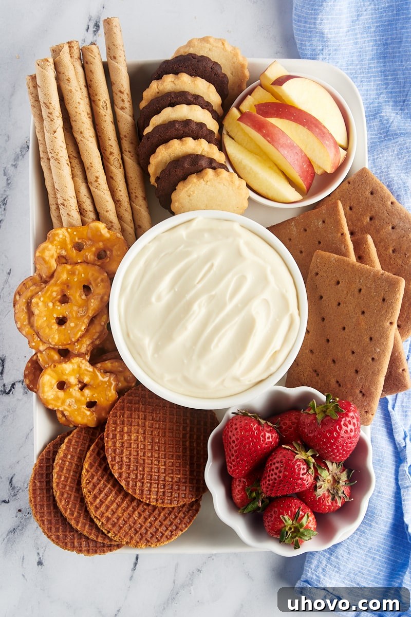 An overhead view of a ready-to-serve cheesecake dip in a decorative bowl, placed on a serving tray surrounded by an inviting assortment of graham crackers, chocolate wafer cookies, and fresh berries like strawberries and blueberries.