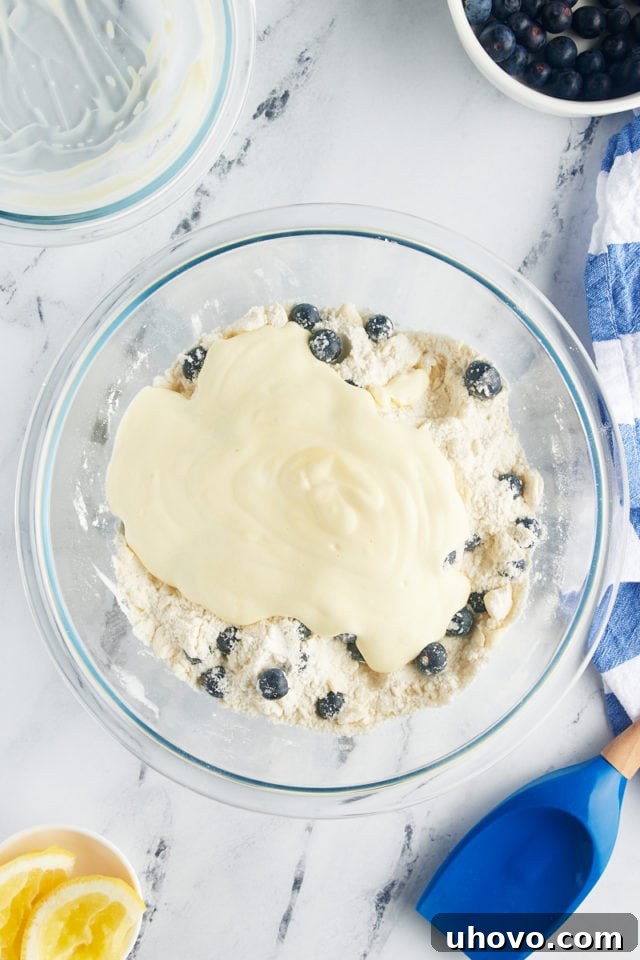 Overhead perspective of the combined wet ingredients (yogurt, egg yolk, lemon juice) being poured into the large glass bowl of dry ingredients for blueberry scones, starting to form the dough.