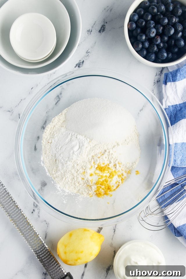 An overhead shot of dry ingredients for blueberry scones – flour, sugar, baking powder, baking soda, salt, and lemon zest – measured out in a glass mixing bowl, ready to be combined.
