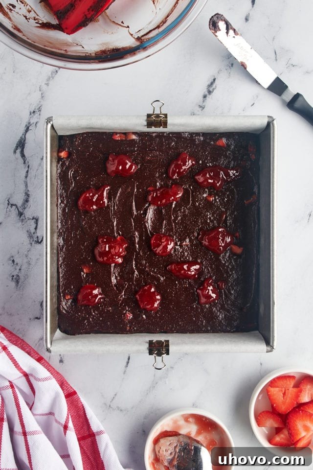 Overhead view of strawberry jam carefully dolloped over the spread brownie batter in the baking pan, ready for swirling.