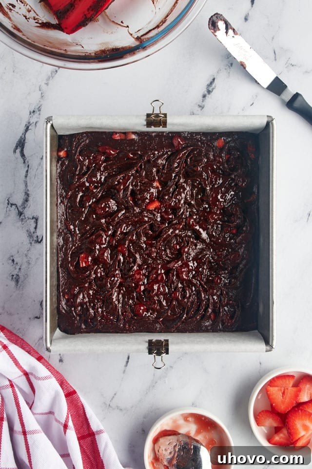Overhead view of brownie batter with strawberry jam swirls and additional fresh strawberries on top in the pan, pre-baking.
