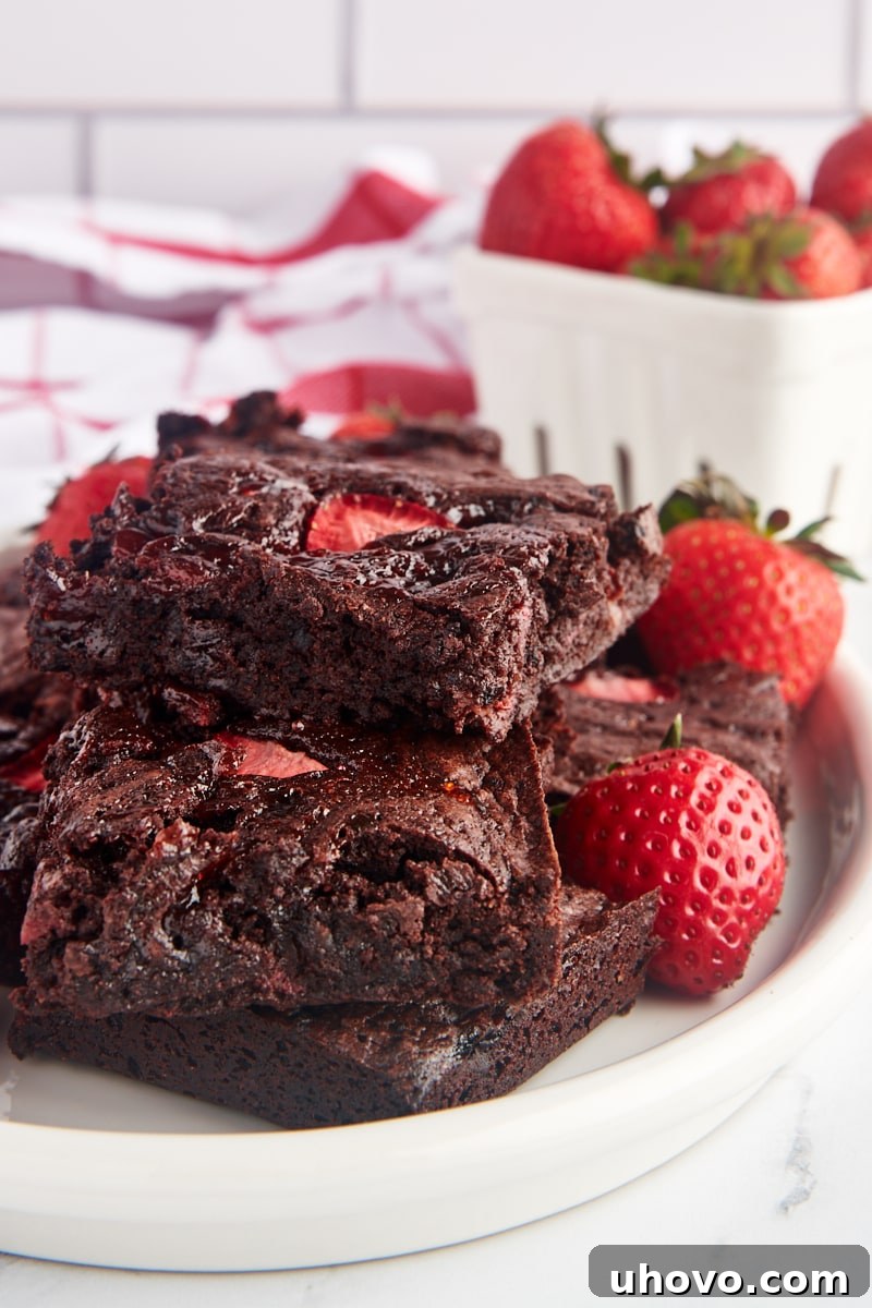 Stack of rich, fudgy strawberry brownies on a white plate, adorned with fresh strawberries, ready to be enjoyed.