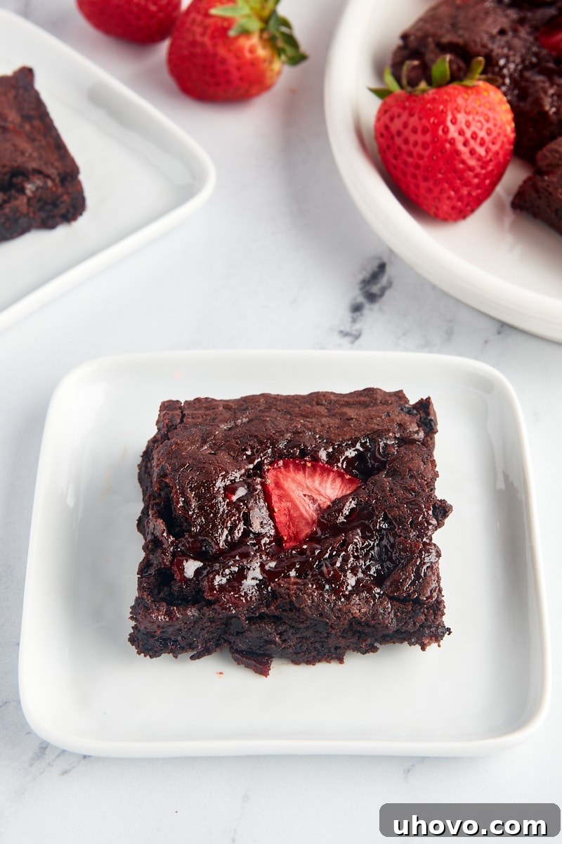 A single fudgy strawberry brownie on a white plate, with additional brownies and fresh berries blurred in the background, inviting you to take a bite.