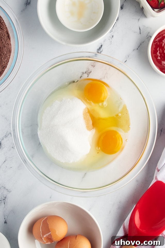 Overhead view of granulated sugar, large eggs, and vanilla extract in a glass mixing bowl, poised for mixing.