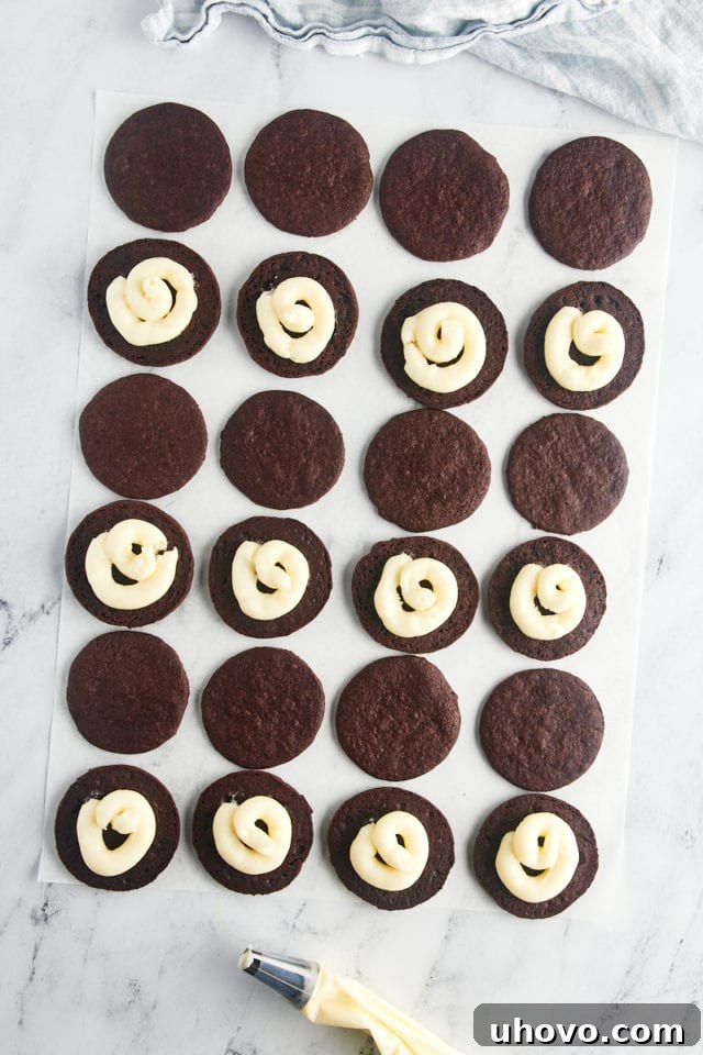 Overhead view of homemade Oreos being meticulously assembled on parchment paper, with creamy white frosting being piped onto chocolate wafers before being topped with another cookie.