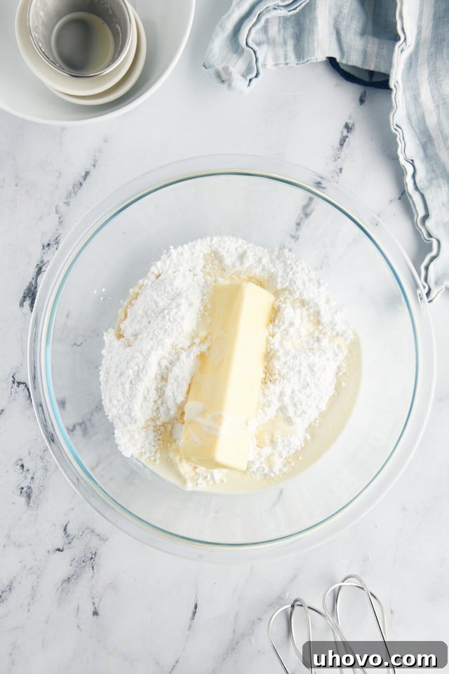 Overhead view of softened butter, powdered sugar, heavy cream, vanilla extract, and a pinch of salt waiting in a clear glass mixing bowl, ready to be transformed into frosting.