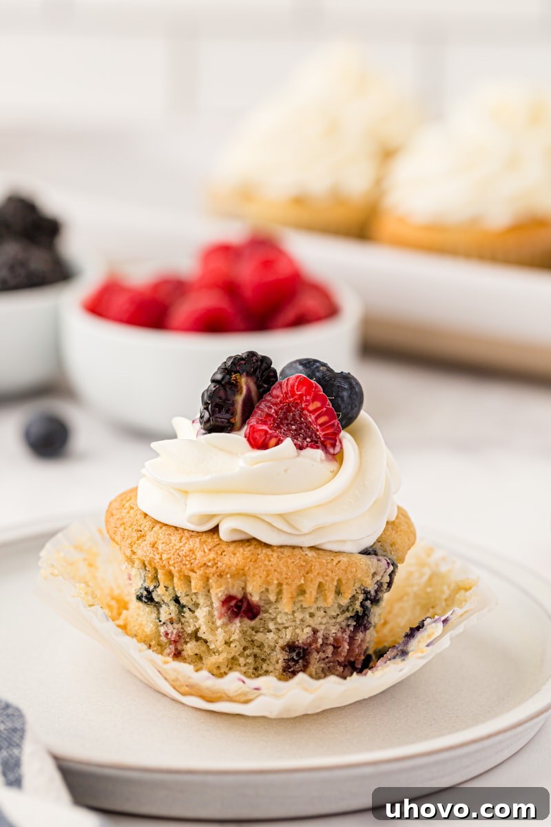 A beautifully frosted berry cupcake on a white plate, with more cupcakes and fresh berries artfully blurred in the background, inviting a delicious tasting.