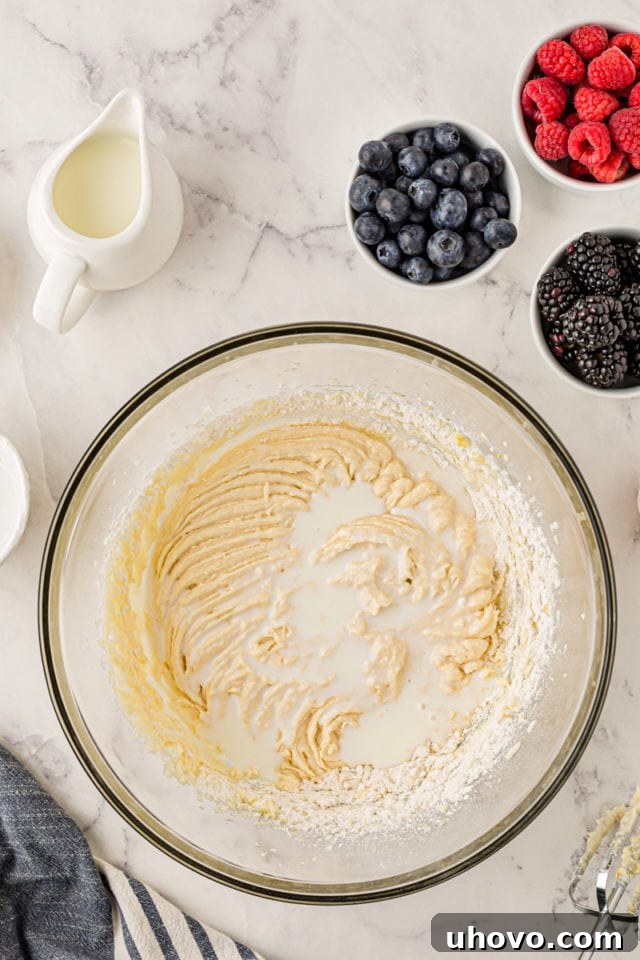 An overhead shot illustrating milk being poured into the partially mixed cupcake batter in a glass bowl, continuing the alternating process with dry ingredients.