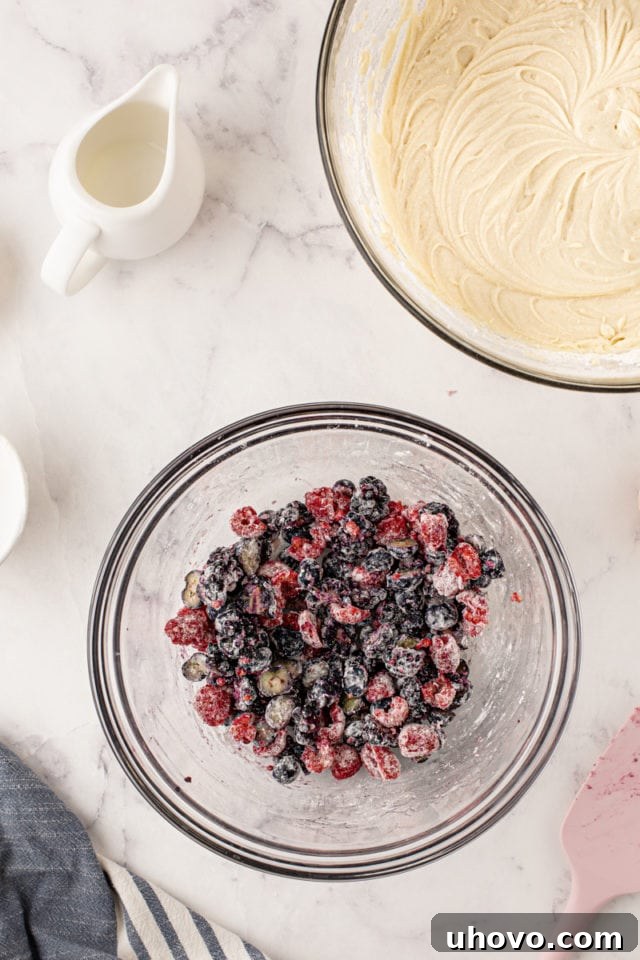 An overhead shot of fresh mixed berries (blueberries, raspberries, blackberries) gently tossed with a light coating of flour in a glass mixing bowl, preventing them from sinking in the batter.