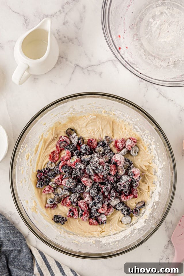 An overhead view of the floured mixed berries being carefully folded into the cupcake batter in a glass mixing bowl, ensuring even distribution without overmixing.