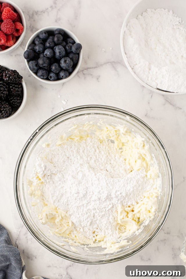 An overhead shot of confectioners' sugar being gradually added to the creamed butter and cream cheese mixture in a glass bowl, for a smooth frosting.