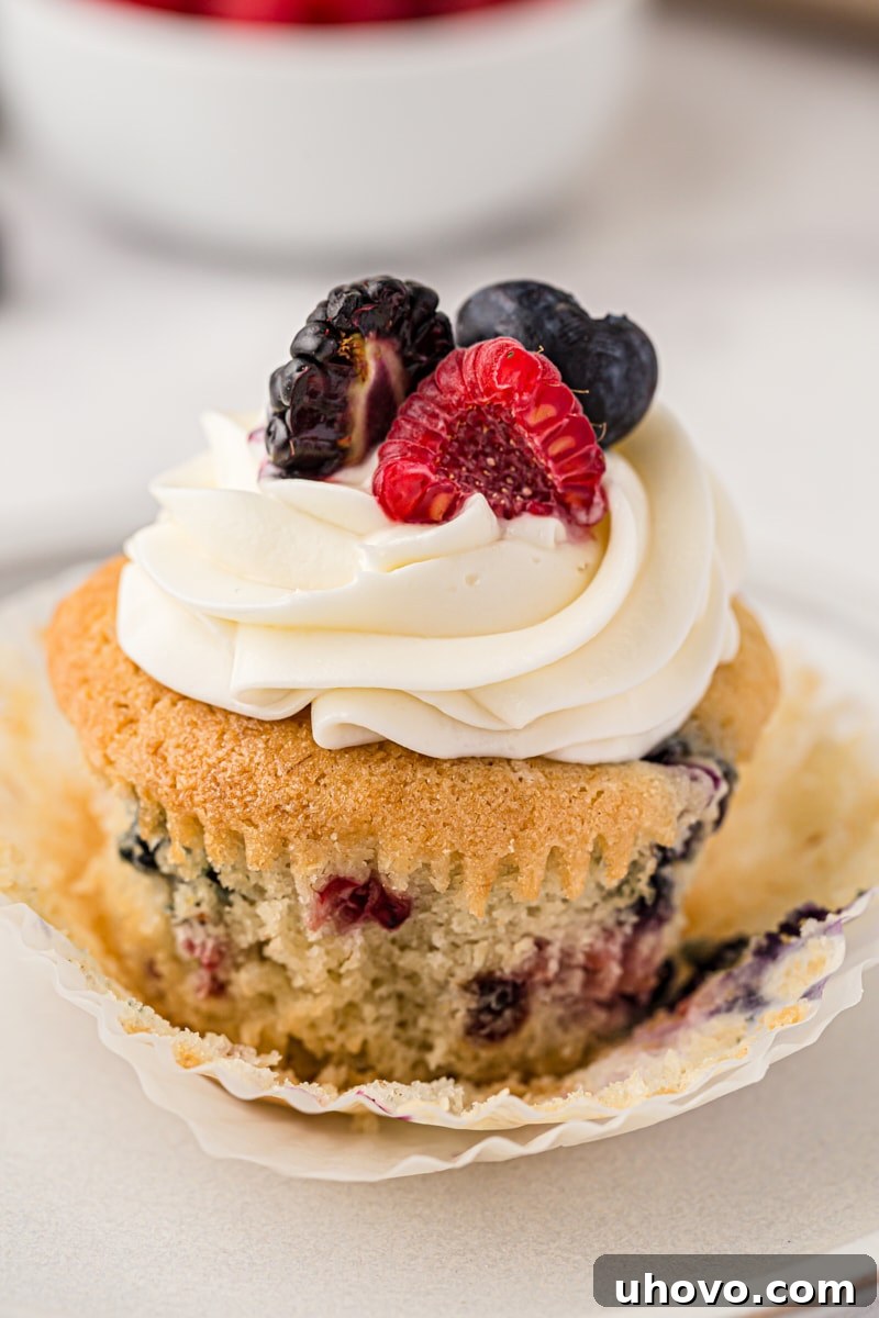 A close-up of a berry cupcake with its paper wrapper partially peeled down, revealing the moist crumb studded with colorful berries, topped with creamy frosting.