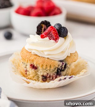 A berry cupcake on a white plate with berries and more cupcakes in the background, highlighting its deliciousness.
