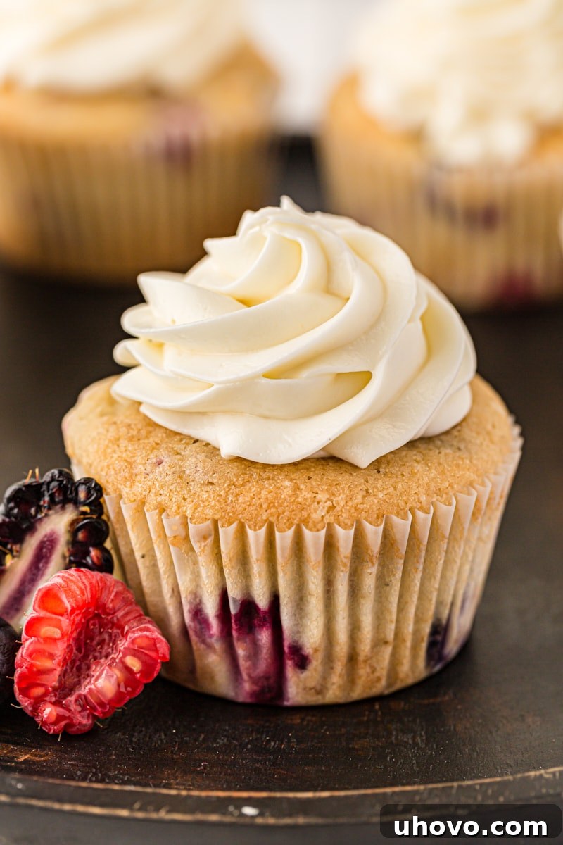 A close-up shot of a single berry cupcake, generously topped with a swirl of rich, creamy cream cheese frosting and a scattering of fresh berries as garnish.