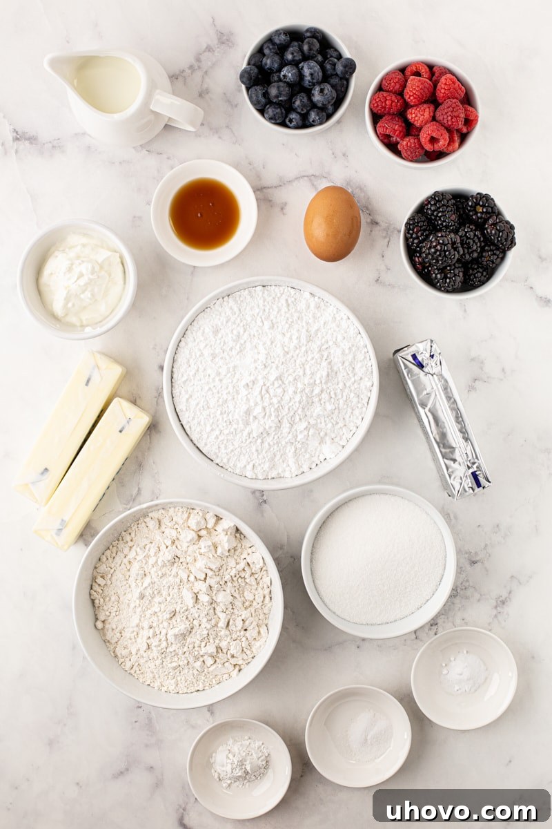 An overhead flat lay of all the essential ingredients laid out on a clean white surface, including flour, sugar, butter, eggs, sour cream, milk, and a colorful assortment of fresh blueberries, raspberries, and blackberries.