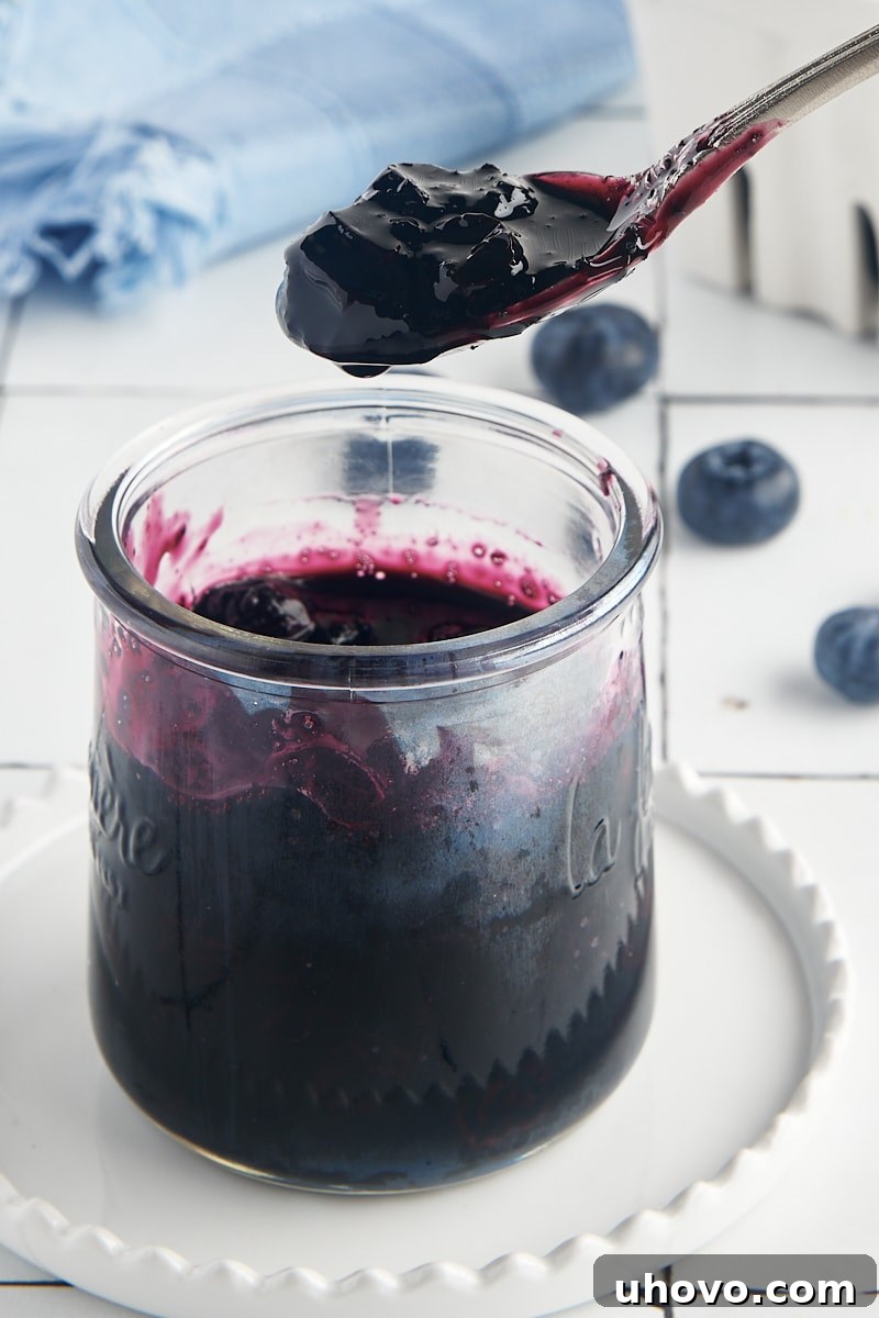 A spoon laden with glistening blueberry compote is held above a glass jar, inviting a taste of its sweet and tangy goodness.