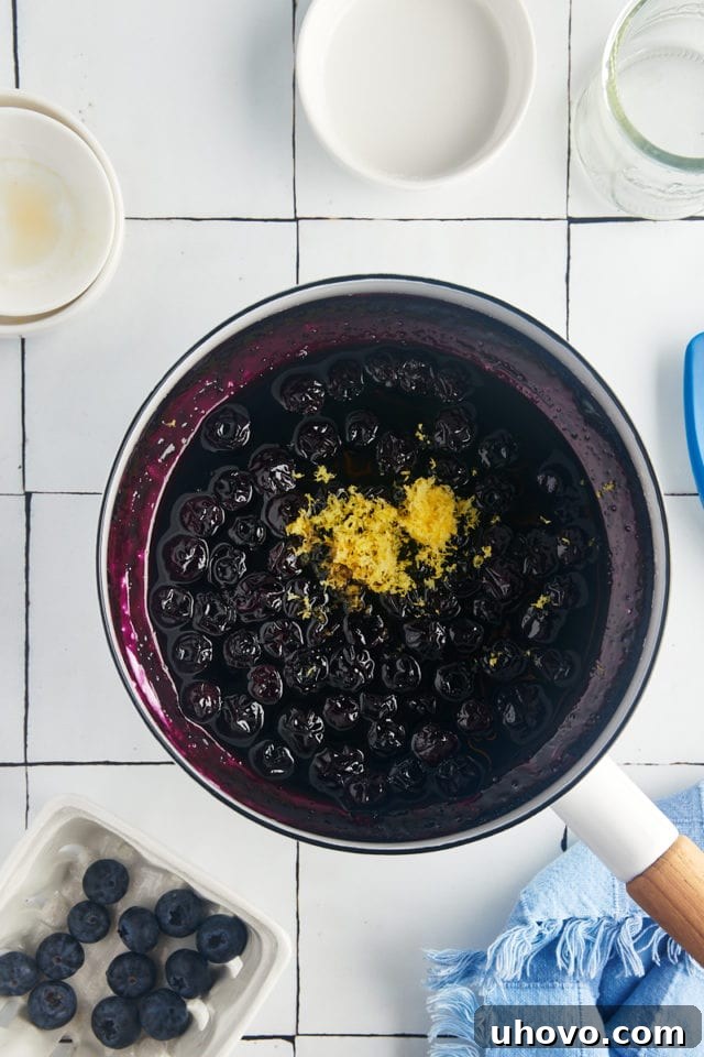 An overhead shot showing vanilla extract and fresh lemon zest being stirred into the cooked blueberry compote in a saucepan.