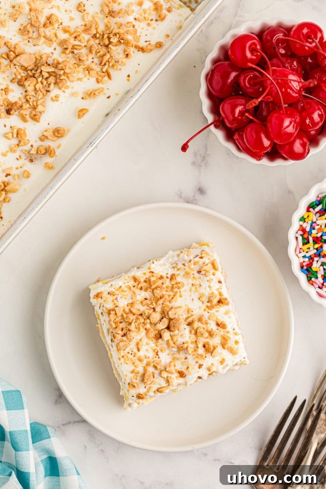 Overhead view of a slice of banana split cake on a plate, garnished with a cherry, with bowls of cherries and sprinkles nearby.