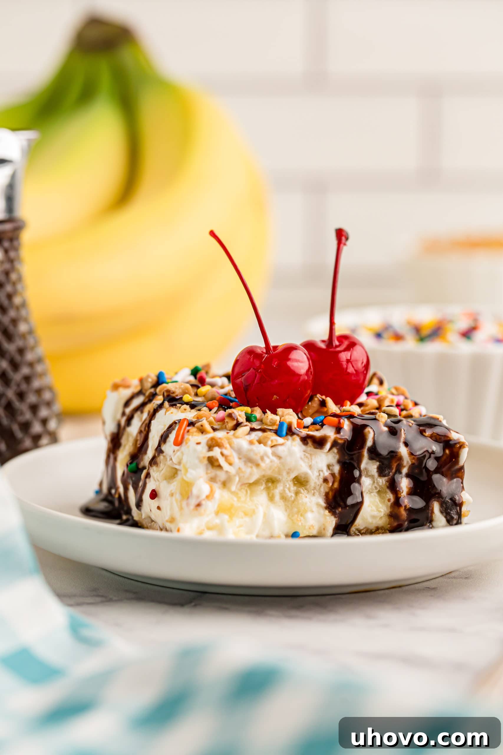 Side view of a generous slice of banana split cake on a plate, perfectly layered and topped with two maraschino cherries.
