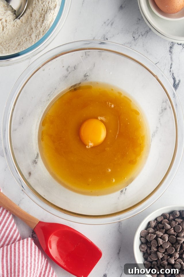 A vibrant overhead view of a large egg and a splash of vanilla extract being added to the wet ingredients for the cookie cake batter, ready for mixing.