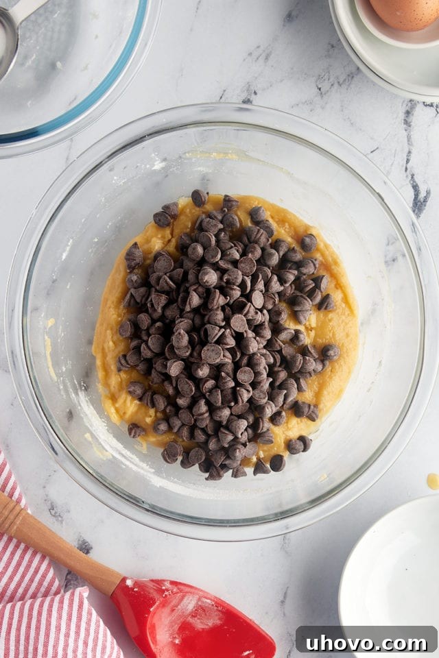 A close-up, overhead view of luscious chocolate chips being generously stirred into the thick, golden cookie batter within a clear glass mixing bowl, promising a burst of flavor.