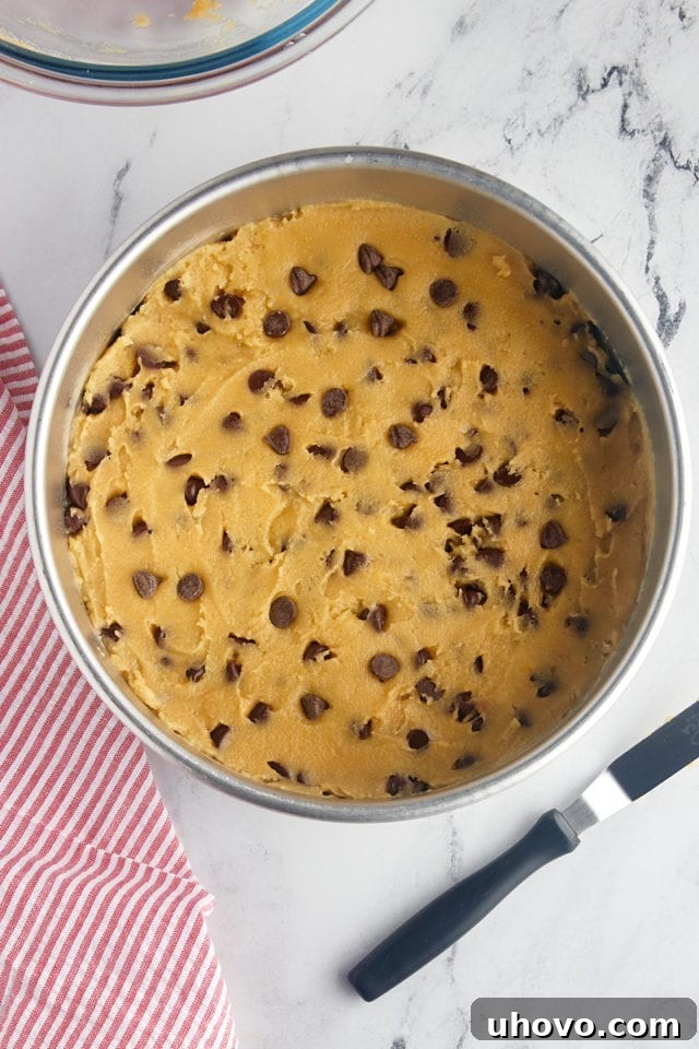 An overhead view of the chocolate chip cookie cake dough expertly spread and smoothed into a round cake pan, ready for its journey into the oven.